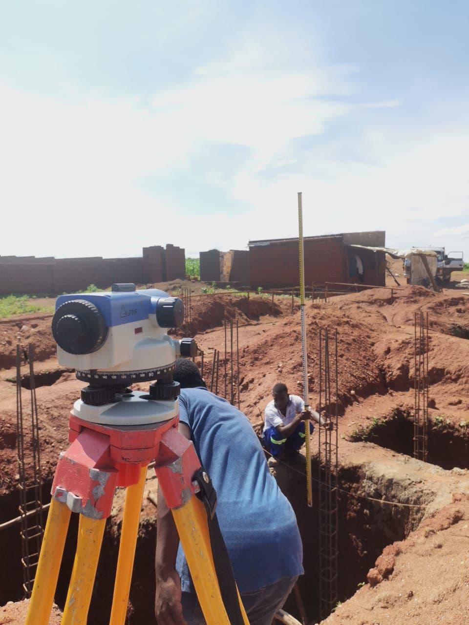 Levelling and setting out. A surveyor's level (dumpy level) was used with a staff to check and confirm excavation depths and levels along the foundation trenches, ensuring the rebar column bases align to the correct height before casting proceeds.