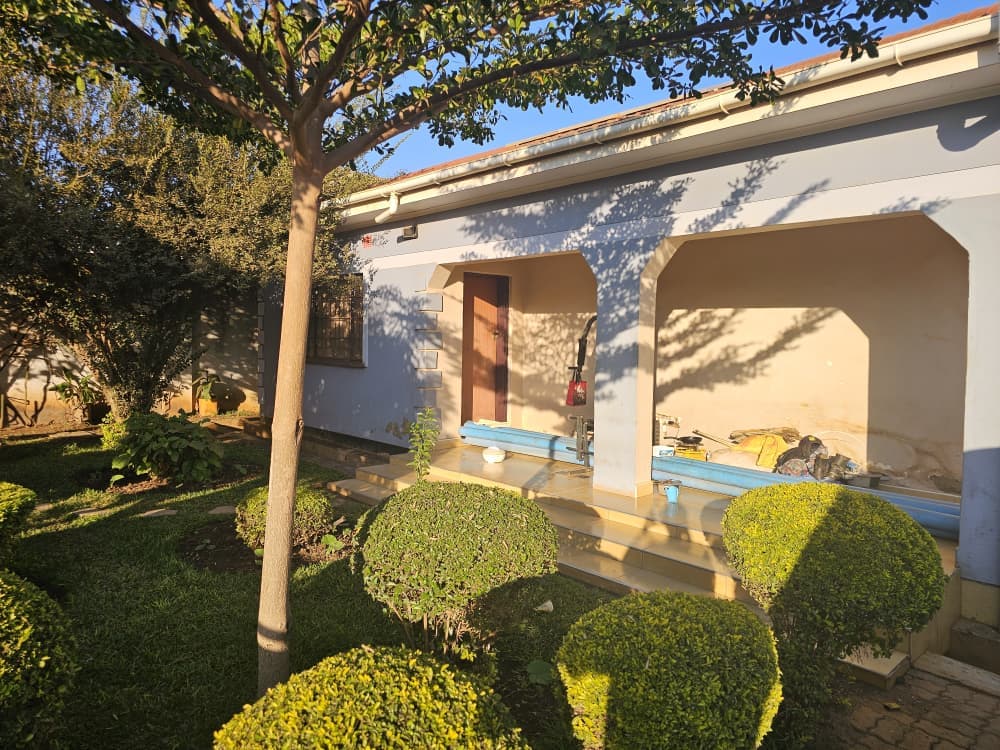 Rendered and painted walls in blue-gray with white trim, textured block-clad columns, tinted aluminum sliding windows and door, tiled veranda flooring, white fascia and gutters, and finished landscaping with a manicured lawn and stepping stones. A clean, modern residential look, completed and ready for occupancy.