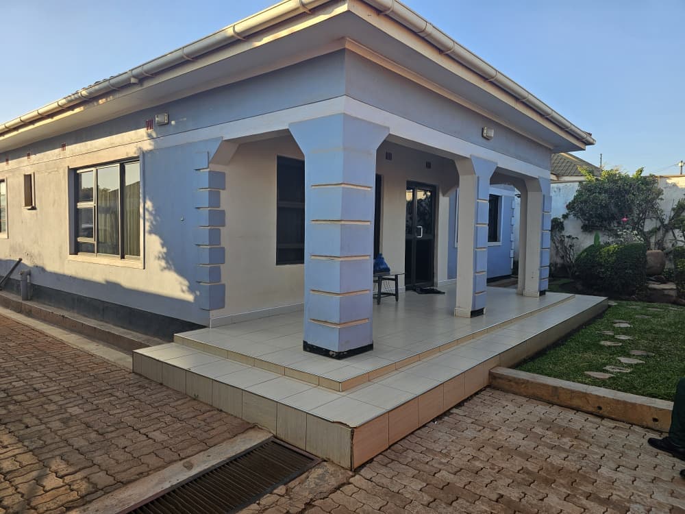 Rendered and painted walls in blue-gray with white trim, textured block-clad columns, tinted aluminum sliding windows and door, tiled veranda flooring, white fascia and gutters, and finished landscaping with a manicured lawn and stepping stones. A clean, modern residential look, completed and ready for occupancy.